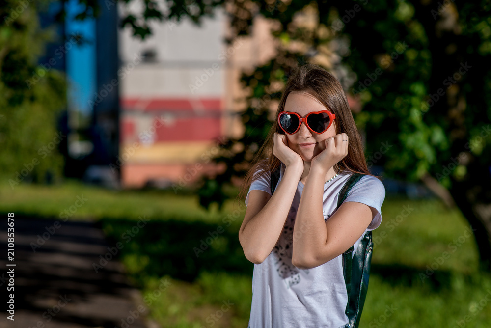 Girl schoolgirl. In the summer after school. Behind the backpack. Sunglasses in shape of the heart. Free space for text. He positively poses for the camera.