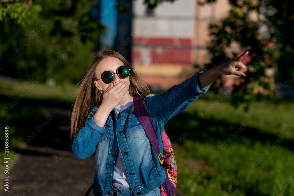 Girl schoolgirl denim jacket, backpack sunglasses. Summer in nature. She covers mouth with palm. Finger points to side. The concept surprise is horror of accident. Emotions of fear misunderstanding. Girl schoolgirl denim jacket, backpack sunglasses. Summer in nature. She covers mouth with palm. Finger points to side. The concept surprise is horror of accident. Emotions of fear misunderstanding.