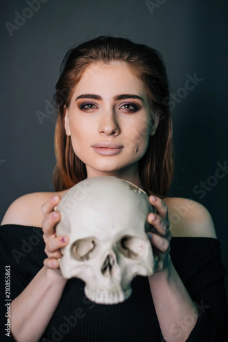 A beautiful girl in black clothes with a skull in her hands. Gothic fashion. A model on a gray background. Studio portrait