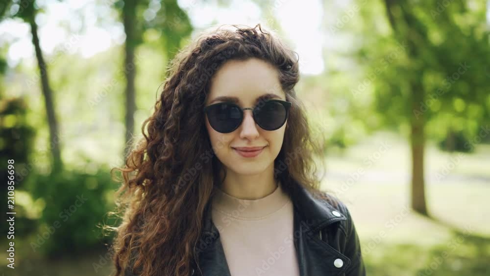 Portrait of attractive woman in sunglasses and trendy leather jacket looking at camera and smiling standing in city park on summer day. People and sunlight concept. Portrait of attractive woman in sunglasses and trendy leather jacket looking at camera and smiling standing in city park on summer day. People and sunlight concept.