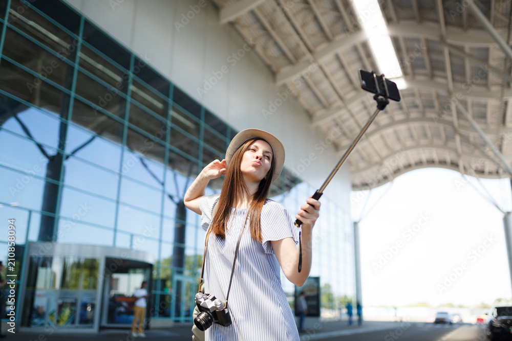 Young funny pretty traveler tourist woman with retro vintage photo camera doing selfie on mobile phone with monopod selfish stick at airport. Passenger traveling abroad on weekend. Air flight concept.