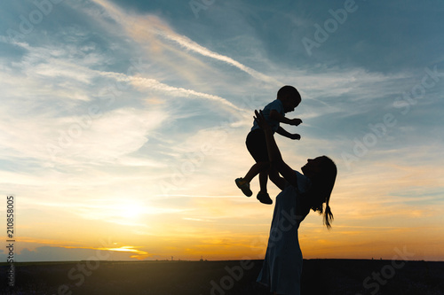 Silhouette of young woman high toss up little cute child baby boy on nature  sunset horizon background. Mother throw up  have fun  play little kid son outdoors. Family day  parents  children concept.