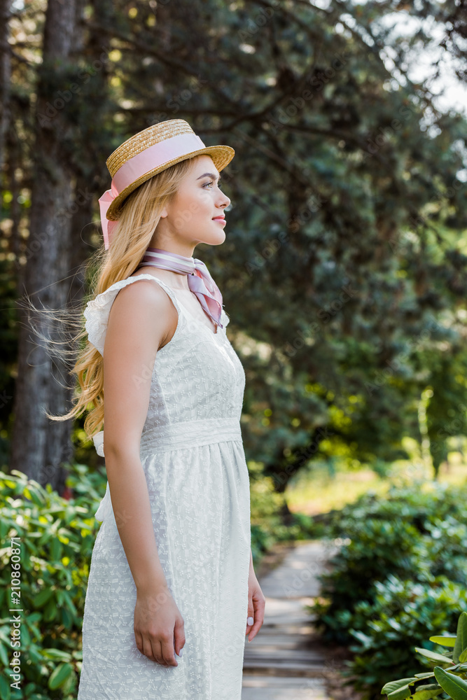 side view of beautiful young woman in white dress and wicker hat smiling and looking away in park