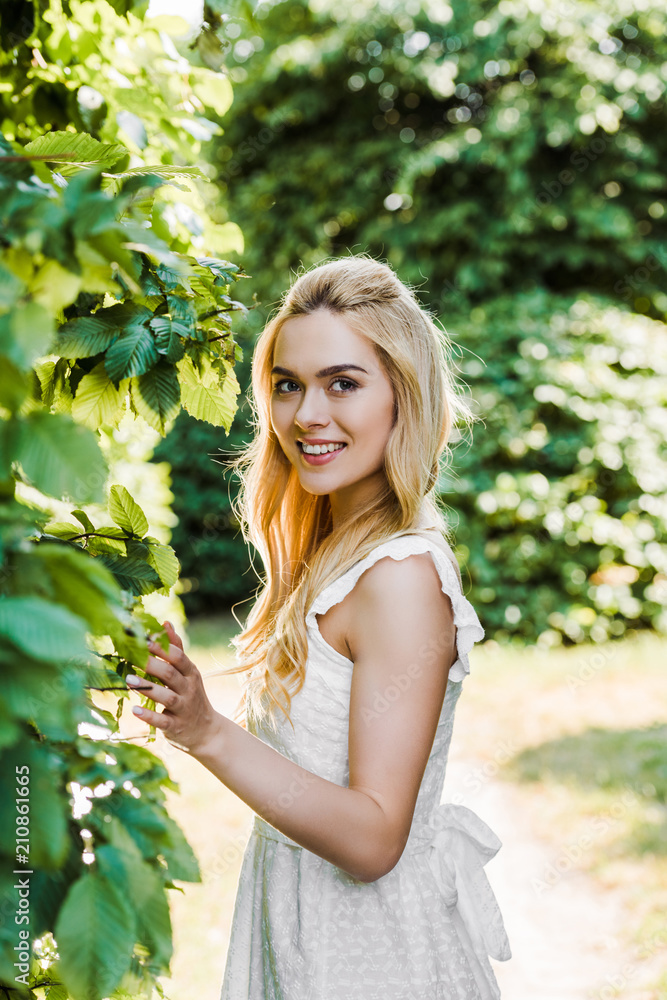 beautiful young blonde woman in white dress touching green leaves and smiling at camera outdoors beautiful young blonde woman in white dress touching green leaves and smiling at camera outdoors