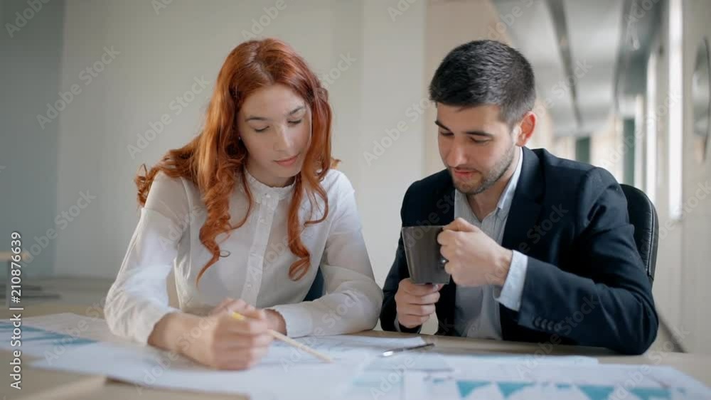 Businesswoman with red hair and male financial analytic together working on investing strategy using printed graphs in the office. Indoors. Straight shot. Slow motion. Businesswoman with red hair and male financial analytic together working on investing strategy using printed graphs in the office. Indoors. Straight shot. Slow motion.
