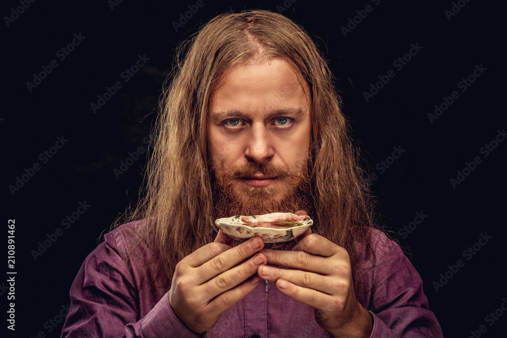 Close-up portrait of a happy redhead hipster male with long hair and full beard dressed in a purple shirt holds a small saucer with tea, looking at a camera. Isolated on dark textured background. Close-up portrait of a happy redhead hipster male with long hair and full beard dressed in a purple shirt holds a small saucer with tea, looking at a camera. Isolated on dark textured background.