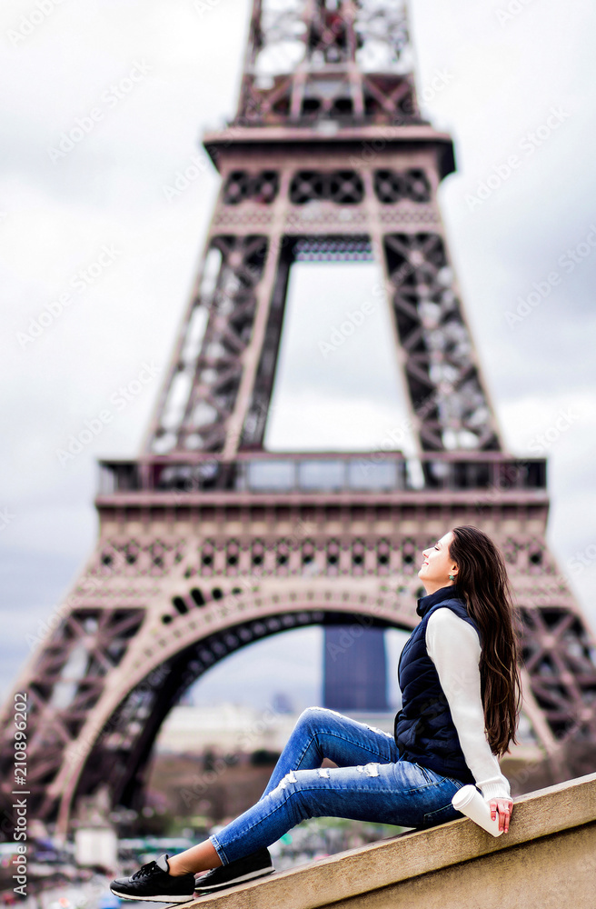 Beautiful young girl in jeans and in a white sweater, holding a coffee on a takeaway on the background of the Eiffel Tower. Concept of a tourist in Paris