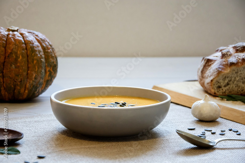 Pumpkin soup with pumpkin seeds. In white bowl, on hessian fabric. With wooden spoon, wholegrain rustic bread on wooden board and whole pumpkin. 