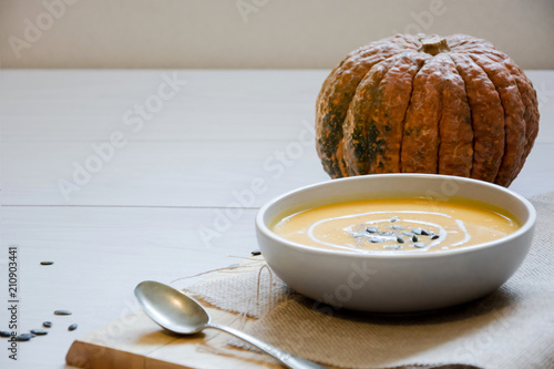 Pumpkin soup with pumpkin seeds. In white bowl, on hessian fabric and wooden board. With silver spoon and whole pumpkin.