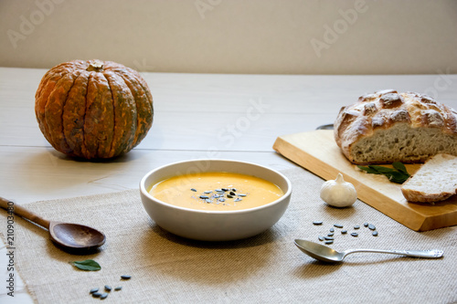 Pumpkin soup with pumpkin seeds. In white bowl, on hessian fabric. With silver spoon, whole pumpkin and rustic bread on a wooden board.