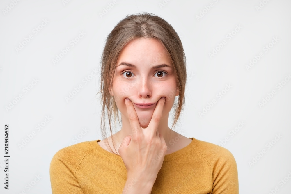 shot of beautiful caucasian teenage girl forcing a smile, holding her fingers at edges of her lips, looking at the camera. Body language