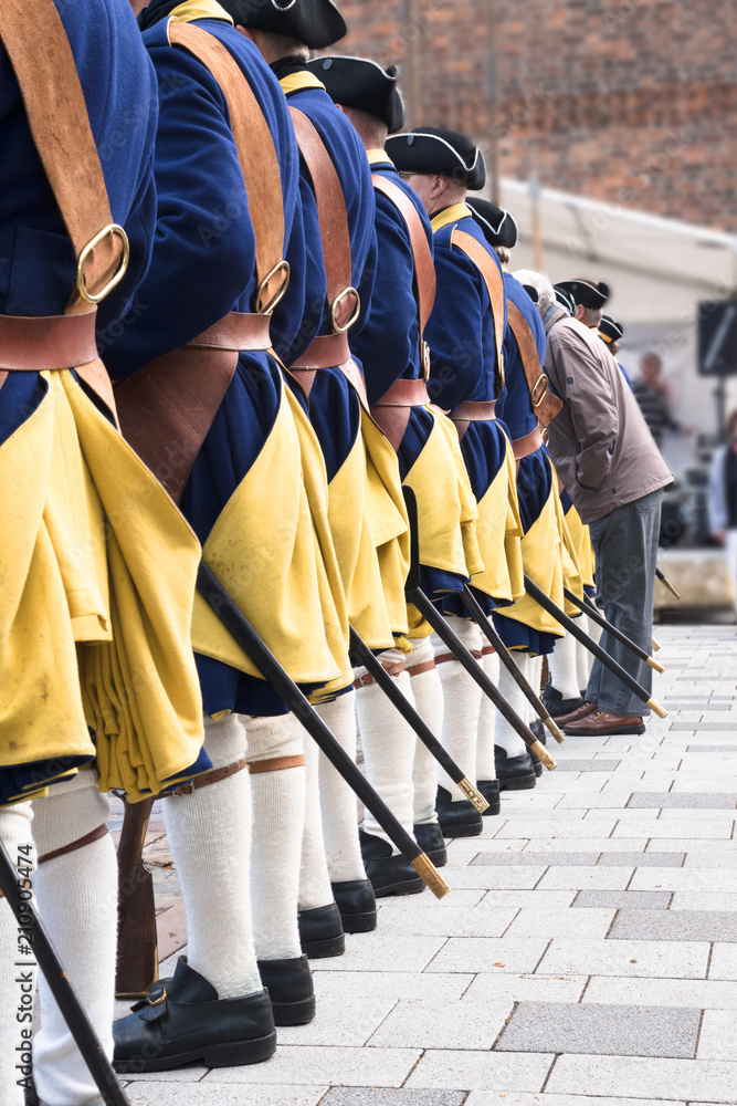 men in a row from behind wear historical swedish soldier uniforms from the XVIII century and one man without costume, vertical