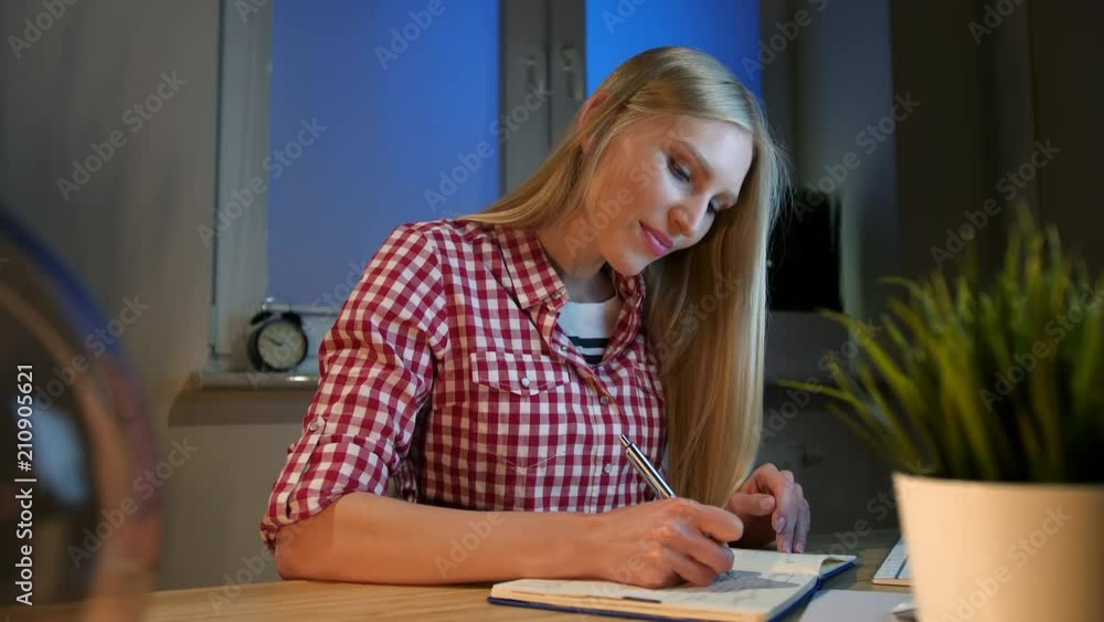 Cheerful female writing in daily planner. Lively young blond woman in casual checkered shirt sitting at wooden desk with notebook holding metal pen and looking at camera smiling at night in dark room. Cheerful female writing in daily planner. Lively young blond woman in casual checkered shirt sitting at wooden desk with notebook holding metal pen and looking at camera smiling at night in dark room.