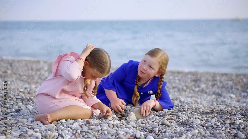 two little girls are sitting on pebble sea coast in daytime, calmly playing on a beach independently two little girls are sitting on pebble sea coast in daytime, calmly playing on a beach independently