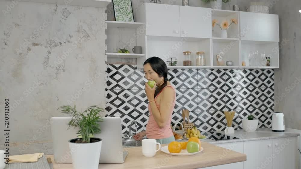 Young Asian woman is eating apple standing in kitchen in apartment, cute brunette is moving, biting green fruit, having time in modern cozy interior with table and laptop. Concept: home time, organic Young Asian woman is eating apple standing in kitchen in apartment, cute brunette is moving, biting green fruit, having time in modern cozy interior with table and laptop. Concept: home time, organic