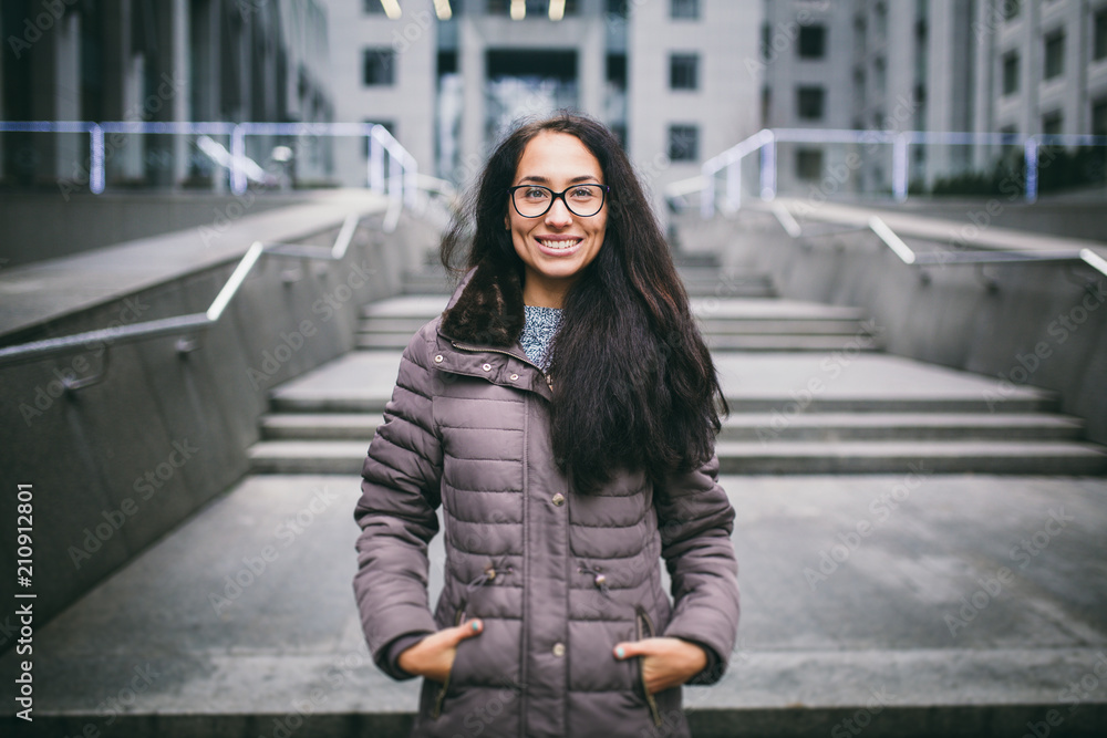 Fototapeta premium Beautiful young woman of European ethnicity with long brunette hair with a toothy smile, wearing glasses and a coat stands against backdrop of a business center in the fall in cloudy cloudy weather