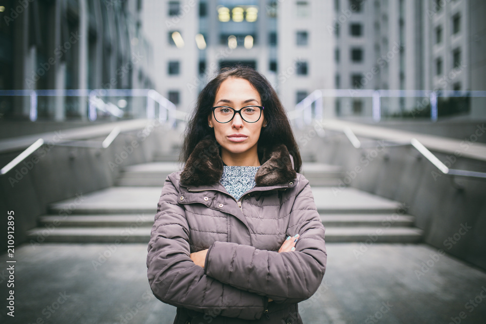 Beautiful young woman of European ethnicity with long brunette hair with grave emotion, wearing glasses and a coat stands against the backdrop of a business center in fall in cloudy cloudy weather Beautiful young woman of European ethnicity with long brunette hair with grave emotion, wearing glasses and a coat stands against the backdrop of a business center in fall in cloudy cloudy weather