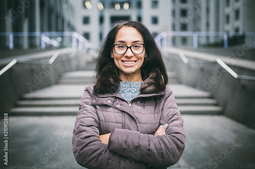 Beautiful young woman of European ethnicity with long brunette hair with a toothy smile, wearing glasses and a coat stands against backdrop of a business center in the fall in cloudy cloudy weather photo
