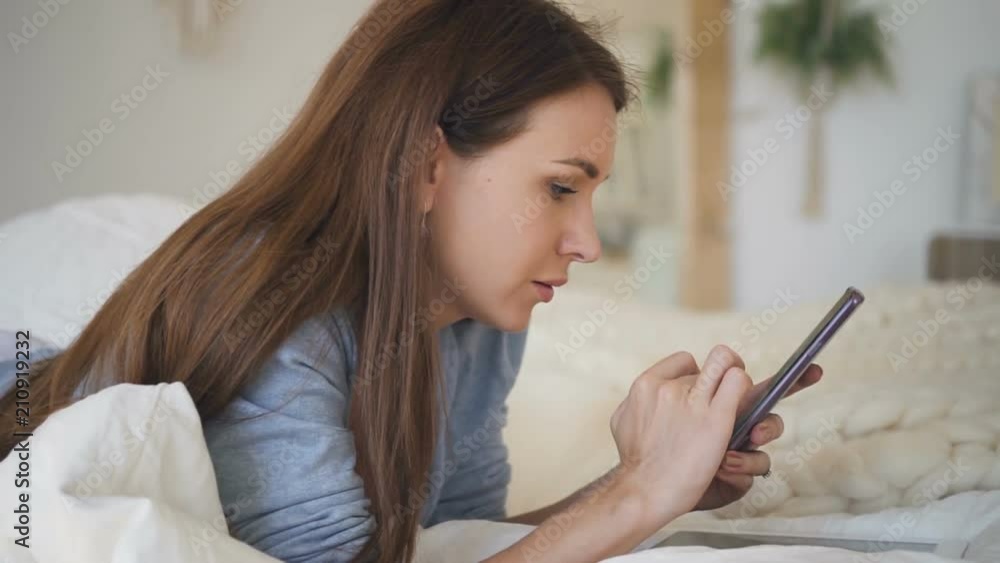 Young woman in blue shirt and with brown hair lying on the bed with white sheets and surfing the internet on her smartphone. Indoors. Portrait. Profile. Young woman in blue shirt and with brown hair lying on the bed with white sheets and surfing the internet on her smartphone. Indoors. Portrait. Profile.