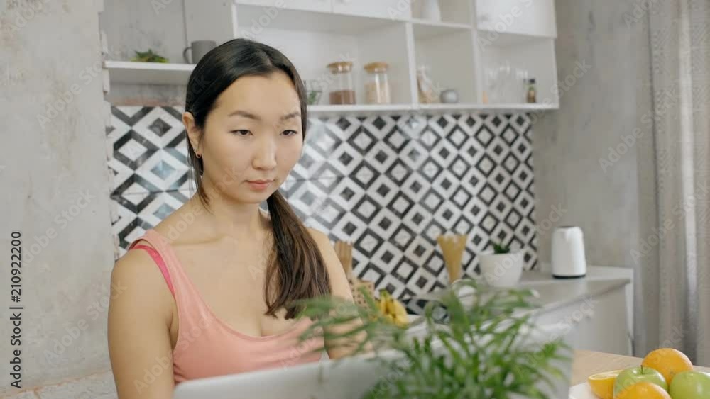 Beautiful asian woman working on her brand new laptop, sitting in the stylish kitchen at home. Indoors. Portrait. Beautiful asian woman working on her brand new laptop, sitting in the stylish kitchen at home. Indoors. Portrait.