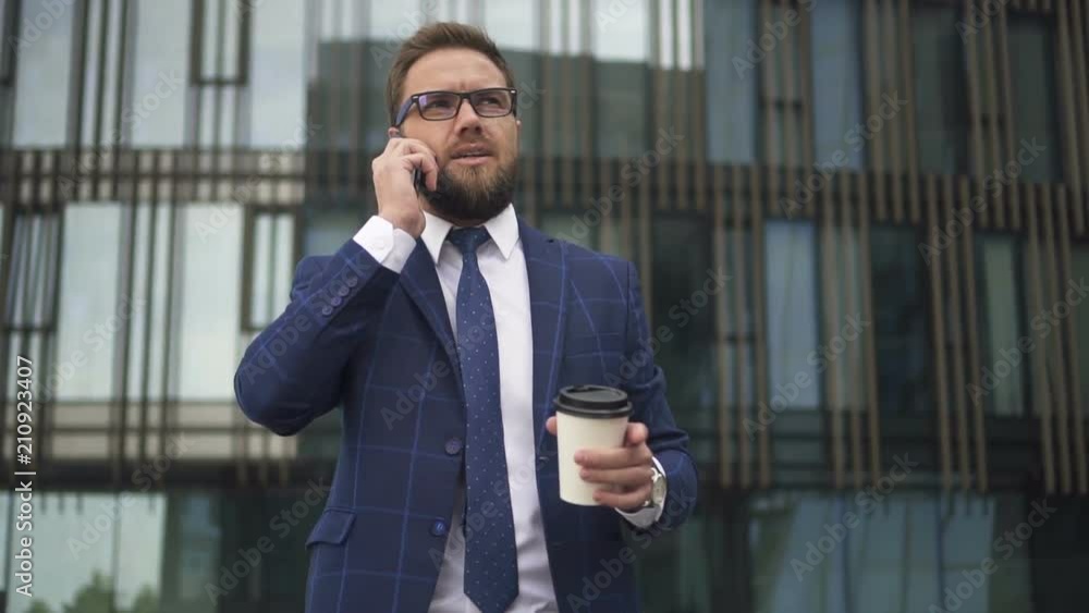Successful businesman talking on phone and holding cup of coffee in his hand standing outside, in business district. Outdoors. Portrait. Successful businesman talking on phone and holding cup of coffee in his hand standing outside, in business district. Outdoors. Portrait.