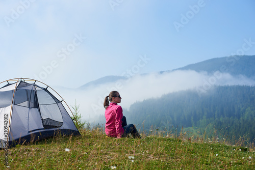 Back view of young long-haired tourist girl in sunglasses sitting on green grass of blooming valley at tourist tent under beautiful clear blue sky enjoying mountains view on bright summer morning.