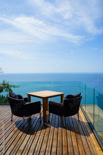 Table and rattan outdoor armchair setting on wooden deck and glass balcony with panoramic Andaman ocean sea view, green tree and blue sky background