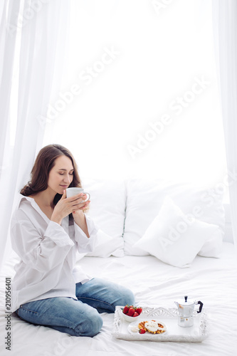 Caucasian young woman sitting on white bed at morning. smelling coffee. breakfast, wafers, strawberry