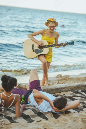 selective focus of young woman playing acoustic guitar for multiethnic friends on sandy beach together