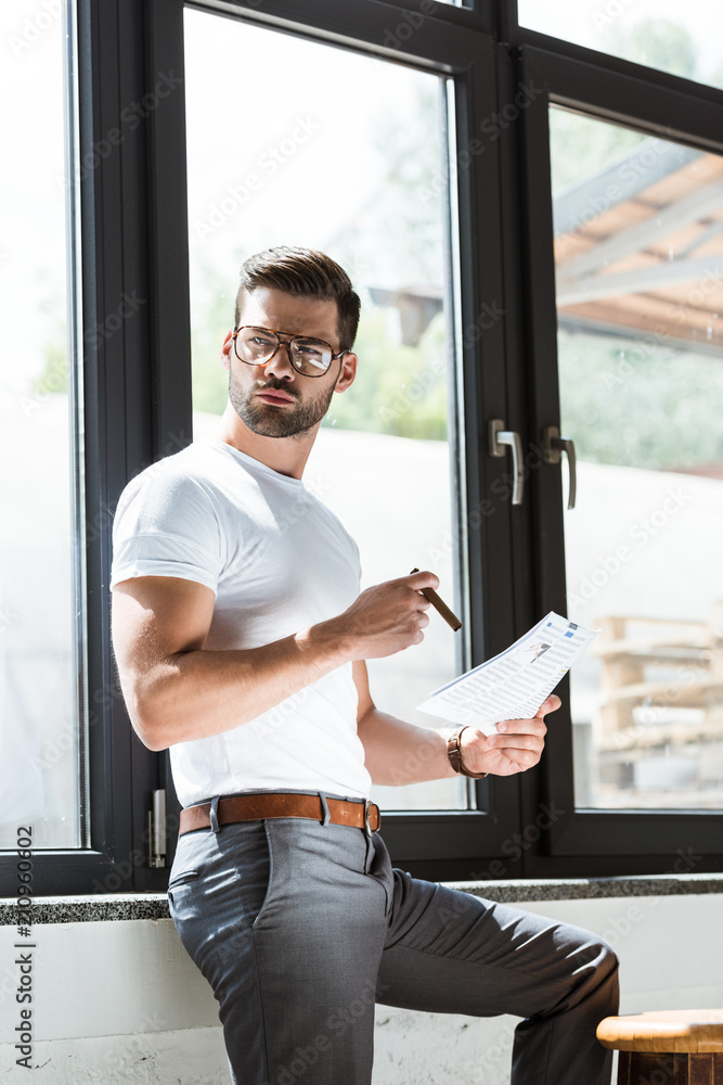 Fashionable confident businessman in glasses smoking cigar while reading business report Fashionable confident businessman in glasses smoking cigar while reading business report