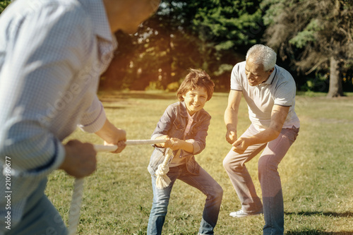 Determined to win. Happy smiling vigorous boy playing rope-pulling with his dad and bending down while grandfather trying to give him a cue
