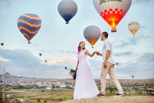 Couple in love stands on background of balloons in Cappadocia. Man and a woman on hill look at a large number of flying balloons. Turkey Cappadocia fairytale scenery of mountains. Wedding on nature