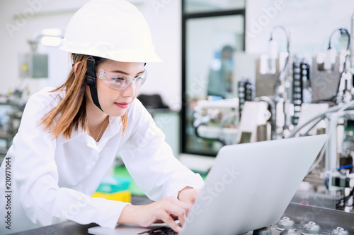 Female industrial engineer wearing hard hat uses laptop at factory.