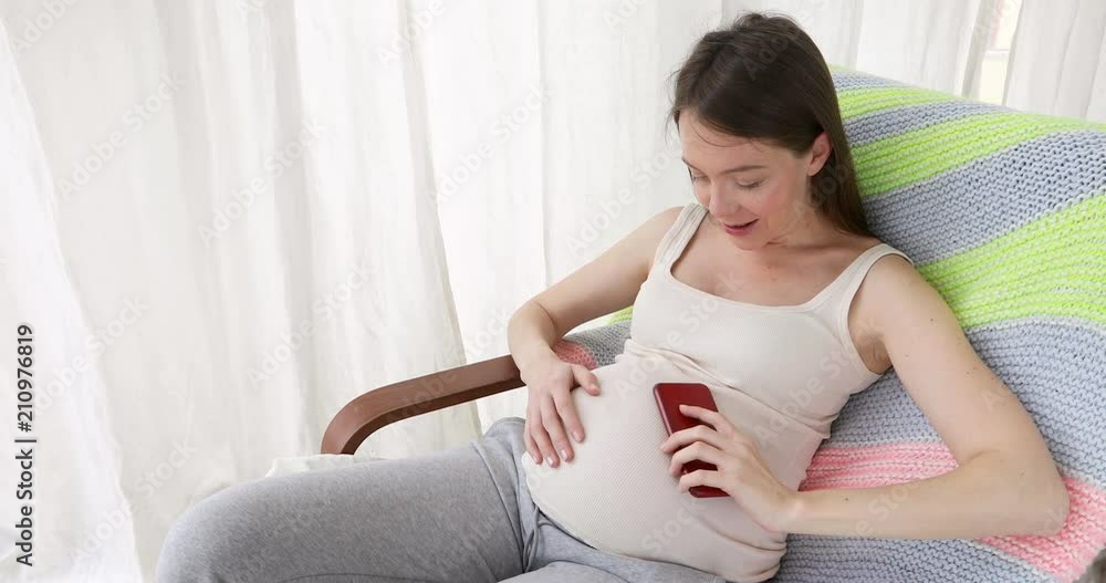 Young pregnant woman sitting in armchair at home and a communicates with a loved one on the phone having funny talk to baby in belly Young pregnant woman sitting in armchair at home and a communicates with a loved one on the phone having funny talk to baby in belly