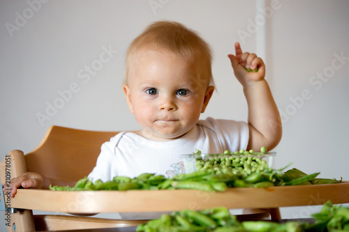 Toddler child, cute boy in white shirt, eating pea at home.