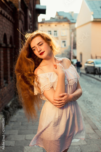 Elegant red haired model with long curly hair wearing white dress posing in evening sunlight © vpavlyuk