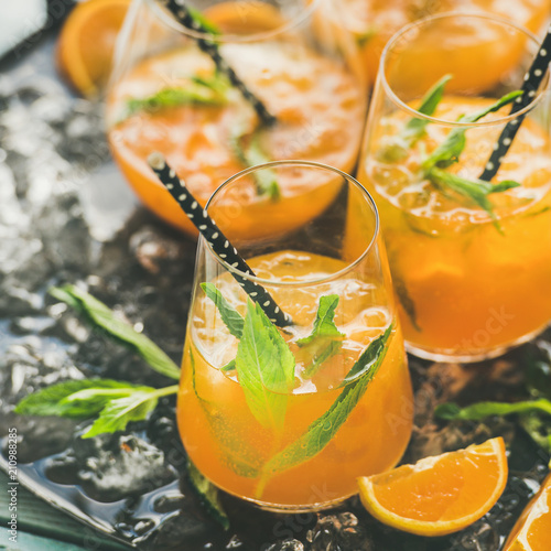 Refreshing cold alcoholic summer citrus cocktail with orange, peppermint and crushed ice in stemless glasses on dark tray over light wooden background, selective focus, square crop