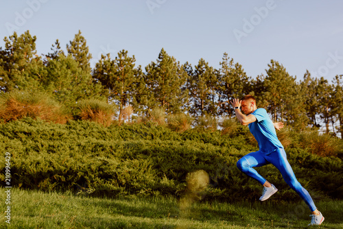 Side view of fit young male jogger exercising on green grass in the park. Handosme athlete running and sprinting outdoor preparing for marathon in mountain. Sport and people concept.