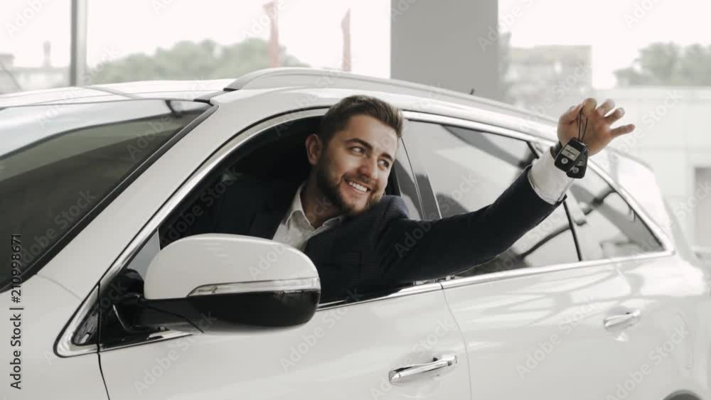 Happy customer is getting out of the car window at the car salon, with the keys of his just purchased car. Happy customer is getting out of the car window at the car salon, with the keys of his just purchased car.