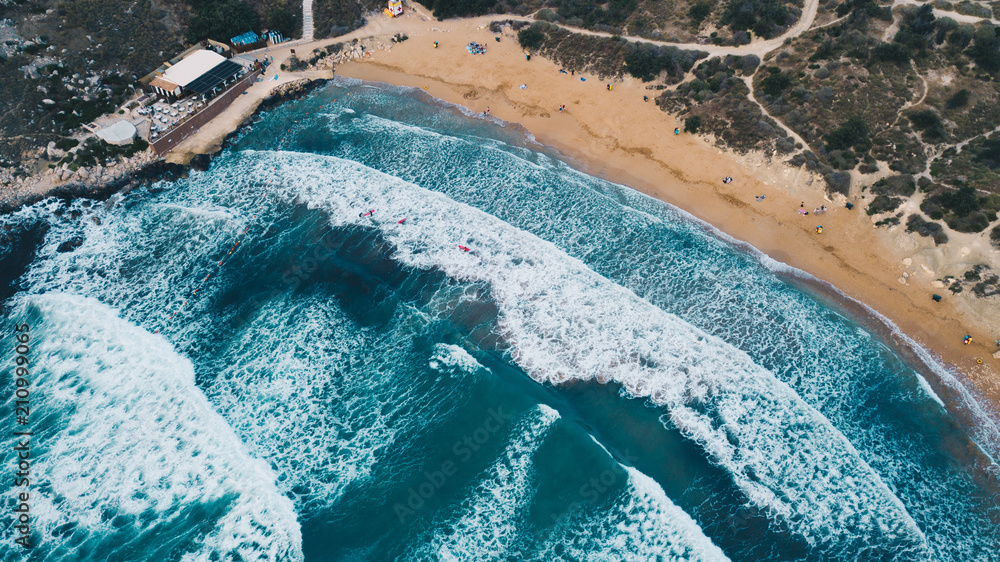 Scenic aerial view of sandy beach and turquoise mediterranean sea with big waves and foam. Surfers with surboats on the waves. Drone view from above. Golden beach, Malta