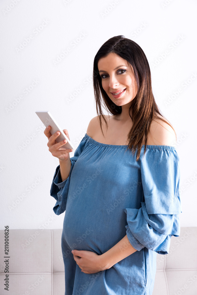 A smiling young pregnant woman standing in a living room in an elegant blue dress and using her smartphone. A smiling young pregnant woman standing in a living room in an elegant blue dress and using her smartphone.