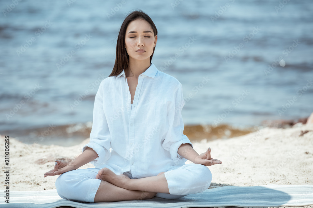 focused woman meditating in ardha padmasana (half lotus pose) on yoga mat by sea