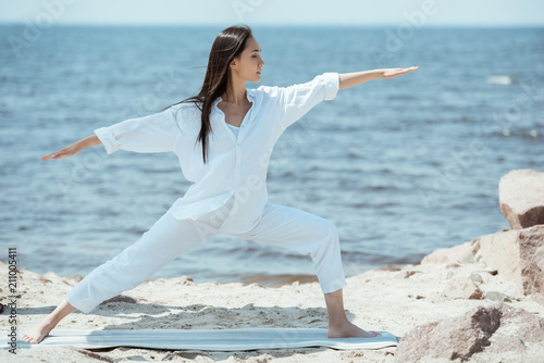 concentrated young asian woman practicing yoga in virabhadrasana (warrior II pose) on mat in front of sea photo