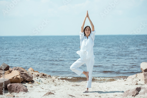 front view of asian woman standing in asana vrikshasana (tree pose) on beach by sea photo