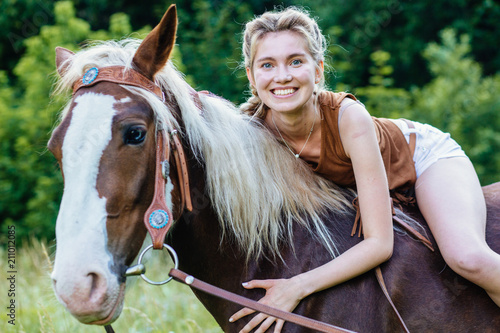 Portrait of happy smiling woman cowgirl, riding a brown horse. Clothed white jeans shorts, brown leather vest. Has slim sport body. Portrait nature. People and animals friendship concept.