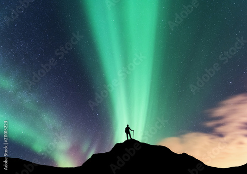 Aurora and silhouette of standing woman on the top of mountain. Lofoten islands, Norway. Aurora borealis and happy girl. Sky with stars and green polar lights. Night landscape with aurora and traveler