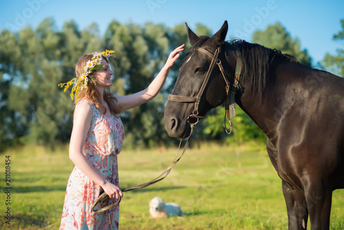 Young woman walking with brown horse on meadow, wearing beautiful dress and midsummer wreath with field flowers