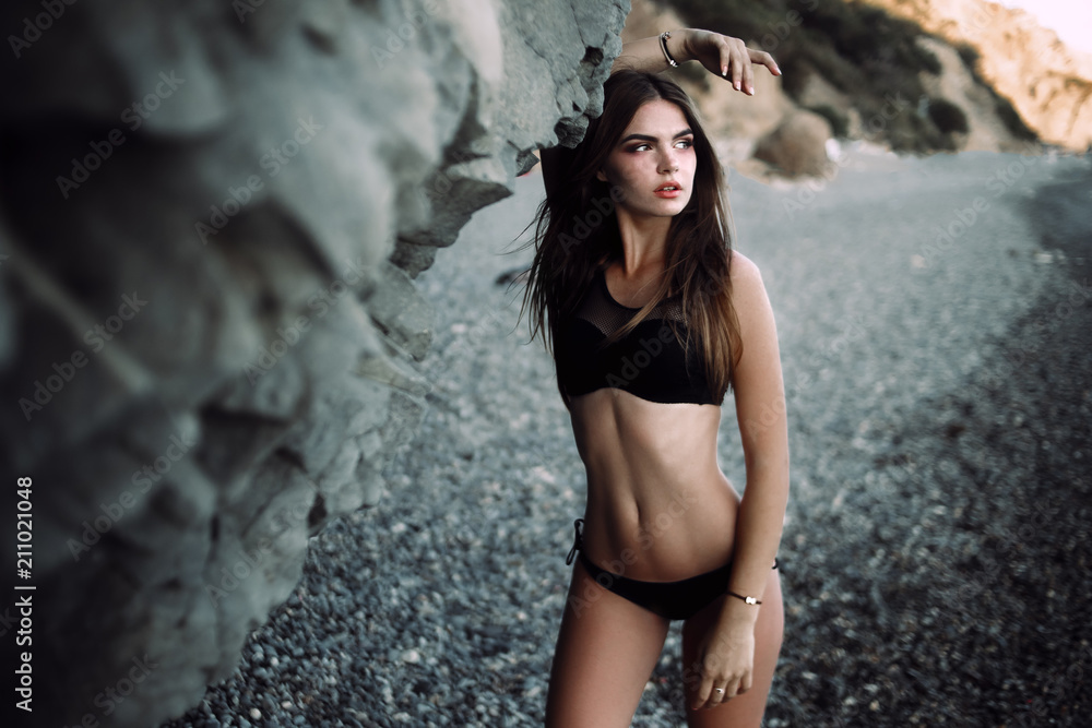 Obraz premium Young sexy woman with beautiful body is posing beside a rocky cliffs on the beach. Horizontal, copy space