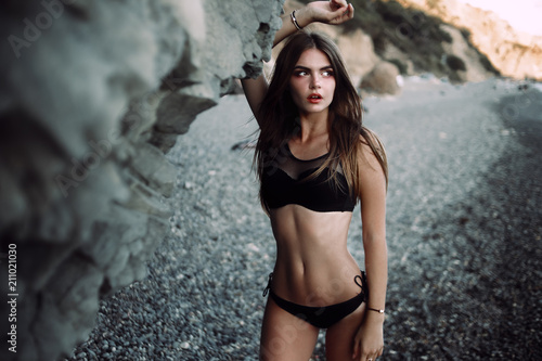 Young sexy woman with beautiful body is posing beside a rocky cliffs on the beach. Horizontal, copy space