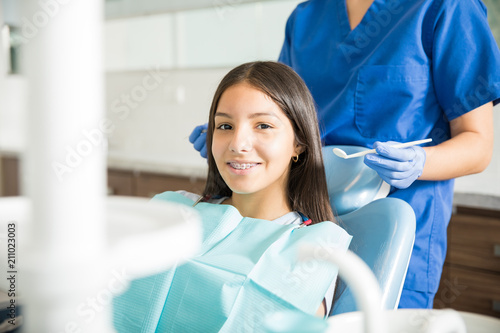 Dentist Standing Behind Smiling Teenage Girl With Braces In Clinic